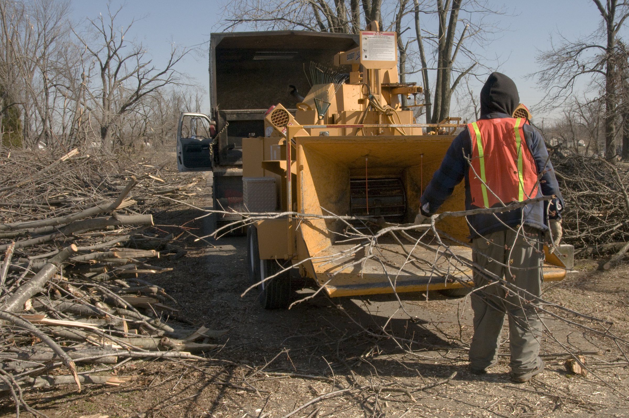 A worker in a safety vest feeds a limb into a towed brush chipper. Storm debris is piled on the ground.