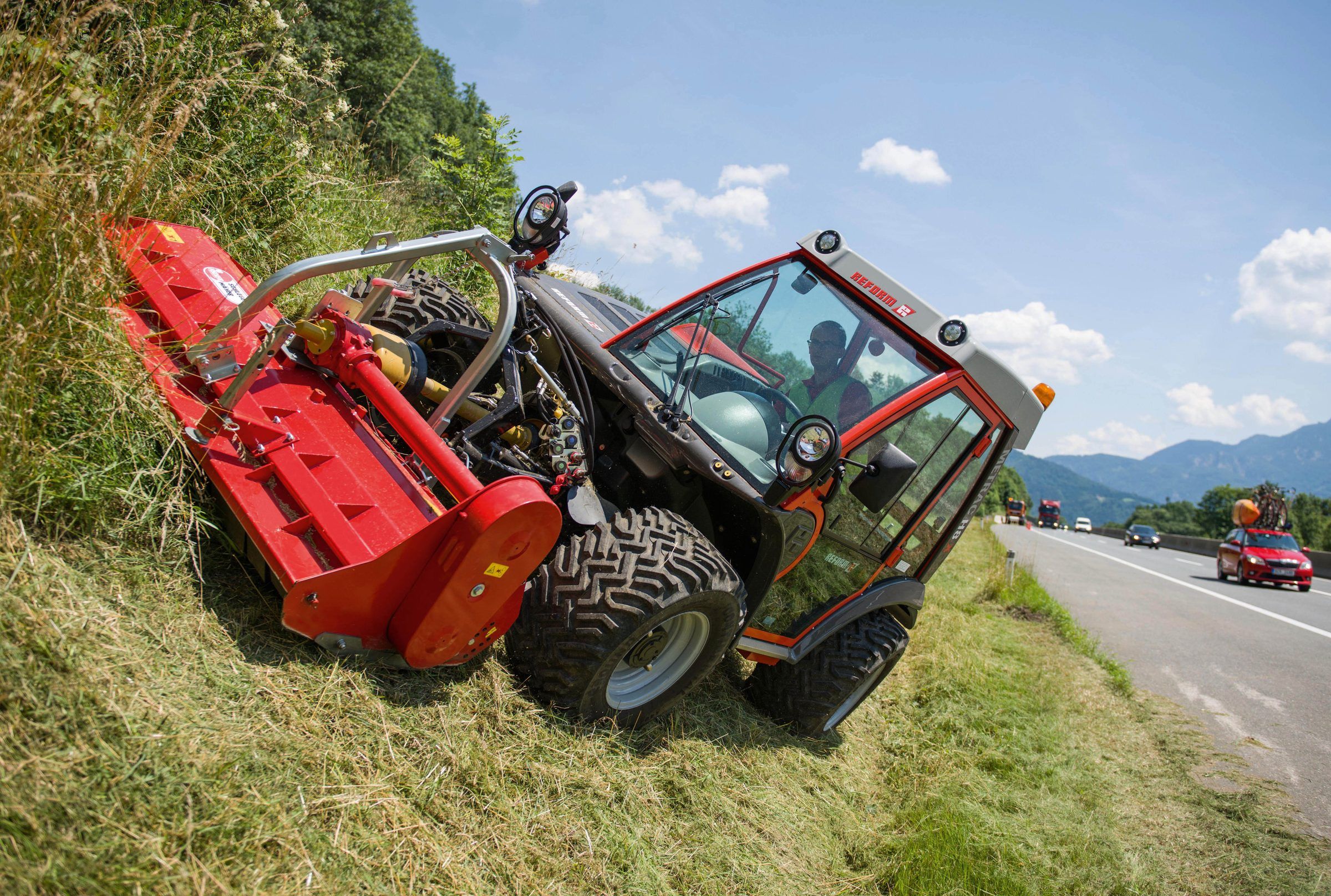 A Reform Metrac articulating slope tractor with a front flail mower cuts tall grass on a steep roadside bank.